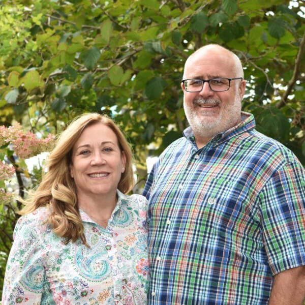 A smiling middle-aged couple standing outdoors, with the man wearing a plaid shirt and glasses, and the woman in a floral blouse, against a backdrop of greenery and pink flowers.