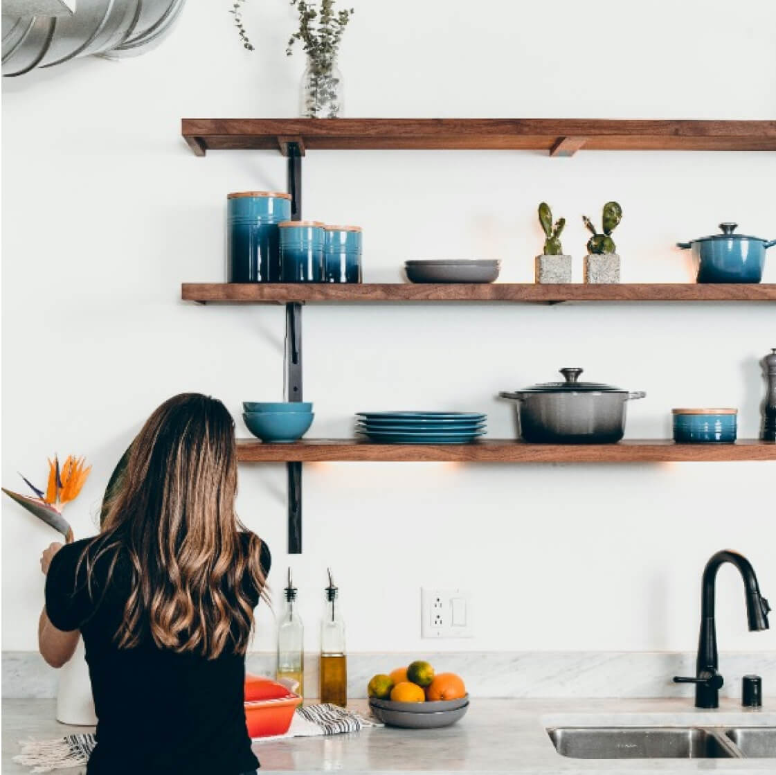 A woman standing in a modern kitchen, reading a cookbook by a countertop with wooden shelves holding kitchenware and plants.