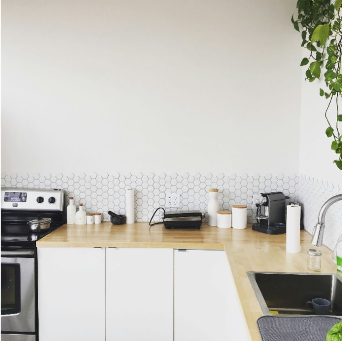 A modern kitchen counter featuring white cabinetry, a stove, coffee machine, and geometric backsplash, with a hanging plant on the left.